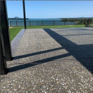 an outside pastio photo showing a bush hammer exposed aggregate concrete floor with a large grass lawn and the sea in the background and a striped fence at the edge of the property beyond the grass