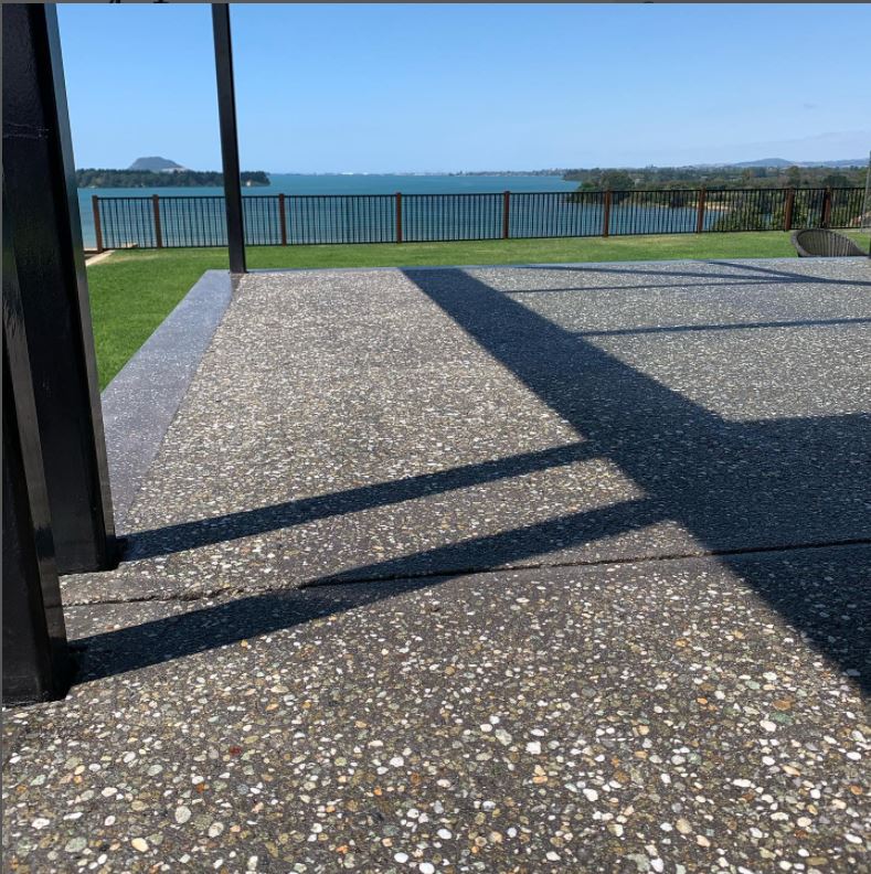 an outside pastio photo showing a bush hammer exposed aggregate concrete floor with a large grass lawn and the sea in the background and a striped fence at the edge of the property beyond the grass