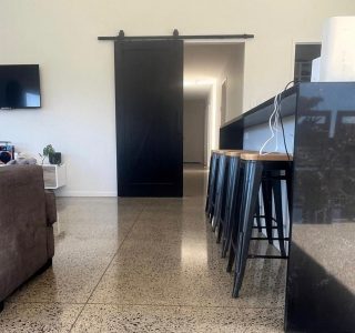 an open plan kitchen diner with the focus on a polished concrete floor, a barn door is in the background painted black and a sofa is to the left, a black kitchen bench to the right with 4 barstools